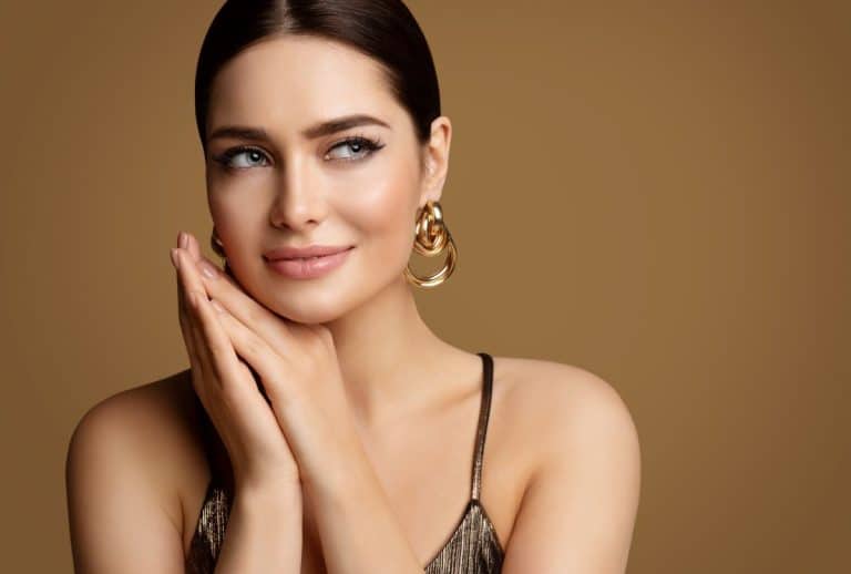Juvederm Filler Candidate Brunette woman with light blue eyes wearing gold earrings. Her hair is in a bun and her hands are placed gently clasped beneath her neck as she looks out of frame to the right. The background is gold.